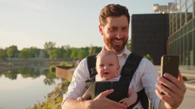 Küçük bir bebek taşıyan, mutlu, genç bir baba. Gün ışığında gölün yakınındaki sokakta selfie çekmek için telefon kullan. Dolayısıyla sırt çantası. Ebeveynler. Gadget. Akıllı telefonmuş. Cep telefonu. Yavaş çekim