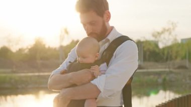 Çekici genç babanın portresi bir çocuk öpücüğü tutuyor ve güneş ışığında kucağına alıyor komik yeni doğmuş bir bebeği kucağına alıyor. Dolayısıyla sırt çantası. Ebeveynler. Yavaş çekim