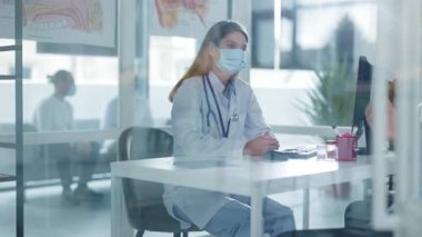 Koruyucu maskeli ve laboratuvar önlüklü kadın doktor klinikteki masada oturuyor. Muayene sırasında erkek hastayla konuşmak. Hastanede. Pandemik koronavirüs tedavisi. Yavaş çekim