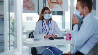 Masasında koruyucu maskeli ve laboratuvar önlüklü bir klinik doktoru oturuyordu. Muayene sırasında erkek hastayla konuşmak. Hastanede. Pandemik koronavirüs tedavisi. Yavaş çekim