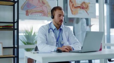 Hastanede, beyaz önlüklü, kulaklıklı genç bir doktor dizüstü bilgisayarla konferans görüşmesi yapıyor. Video sohbetinde hastaya danışmanlık yapıyorum. Yavaş çekim