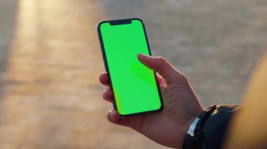 NEW YORK - 19 Aralık 2020: Şehrin arka planında dikey yeşil ekran güneş ışığı yolu olan dokunmatik telefonu kullanan erkek eli bulanıklaştı. İnternet krom açık hava cep telefonu akıllı telefonları kayıyor. Yavaş çekim