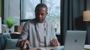 Heyecanlı Afrikalı adamın portresi iş yerinde dizüstü bilgisayarla oturuyor, akıllı telefonu alıyor ve iyi haberlerle mesajları kontrol ediyor. Serbest İşçi 'nin günlük hayatı. İçeride. Ev