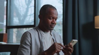 Şaşırmış Afrikalı adamın yatay çekimi akıllı telefon ekranında haber yayını yapıyor. Neşeli iş adamı sosyal medyayı kullanıyor ve evde haberleri okuyor. Kapalı