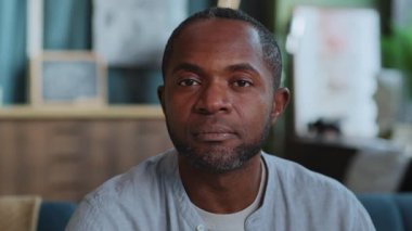 Oturma odasında kameraya odaklanmış Afro-Amerikan bir adamın yatay yakın plan portresi. Modern dairenin bulanık arka planında kendine güvenen bir serbest yazar. Gündüz vakti. İç mekan. Kapalı