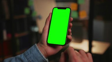 Modern akıllı telefonun erkek ellerindeki görüntüsü akşam ofisin bulanık arka planında izole edilmiş. Yeşil ekranlı bir cihaz kullanan, tanınmayan bir adamın yakın çekim görüntüsü. Krom anahtar kavramı. Kapalı