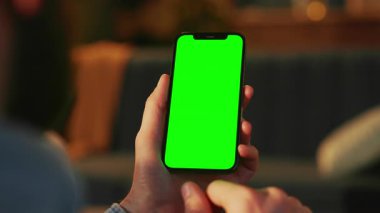 Kanepeli oturma odasının bulanık arka planında yeşil ekranlı akıllı telefonu tutan adamlar. Tanımlanamayan sakallı hippi haber bülteni. Kapalı