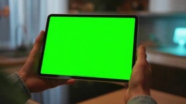 İzole edilmiş sol elin yeşil ekranlı dijital tableti tutuş görüntüsü. Tanımlanamayan beyaz adam ekrana dokunuyor, kaydırıyor, dokunuyor, parmağıyla mutfağı işaret ediyor.