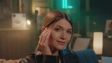 Saça dokunan ve evdeki kanepede oturan kameraya bakan gülümseyen genç bir kadının portresi. Gece lambası. Rahatla.