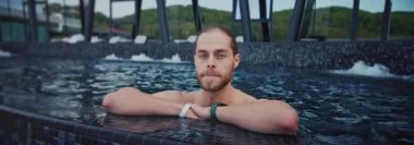 Yüzme havuzunda dikilen adam kameraya bakıyor. Açık havada. Rahatlama zamanı. Tatil