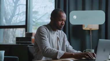 Odaklanmış bir adam rahat bir masada dizüstü bilgisayarı ve sıcak bir kahveyle oturuyor, iş ve özel hayatını dengelerken cep telefonuna bakıyor.