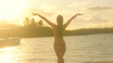 Bikinili bir kadın, kolları neşe içinde ışıldayan suda durarak huzurlu bir günbatımının tadını çıkarır. Altın renkleri sahneyi güzelleştirirken yakınlarda bir tekne süzülüyor.