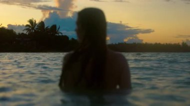 Sakin bir okyanus gün batımı, sığ sularda bikinili bir kadının saçlarının aktığını gösteriyor. Güneş batarken, sakin bir yüzüşün tadını çıkarıyor, yazları dinlenirken yakalıyor.