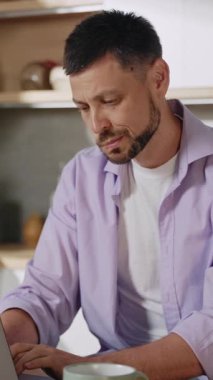 Bir adam evinde dizüstü bilgisayarıyla düşünceli bir şekilde çalışıyor ve modern ve şık bir mutfakta güzel bir kahvenin tadını çıkarıyor.