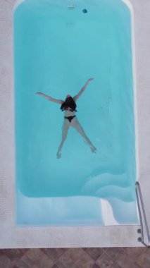 Sakin bir yaz sahnesinde, bikinili bir kadın zarif bir şekilde süzülürken bir kişi köpüklü bir havuzun kenarında görülüyor. Kamera havuzu ve şezlongları yakalıyor, rahatlamak için mükemmel.