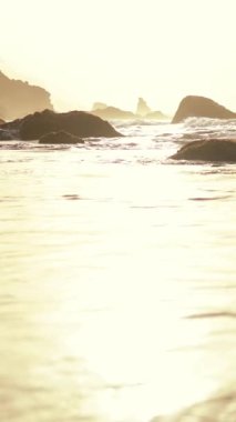 Sakin ve huzurlu bir sahil sahnesi, yumuşak, sıcak güneş ışığı altında engebeli kayaların yanında parıldayan suları yakalıyor.