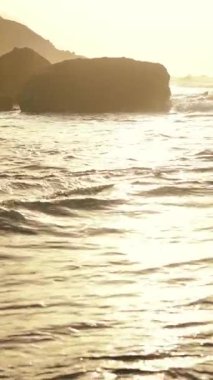 Güzel, sakin bir günbatımı manzaralı kayalık bir kıyı şeridinin yakınındaki yumuşak okyanus dalgalarına yansıyor.