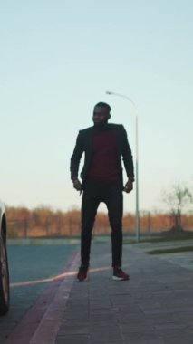 Videoda takım elbiseli iyi giyimli ve kırmızı kazaklı bir adamın yoğun bir kaldırımda yürüdüğü görülüyor. Pırıl pırıl beyaz bir arabanın yanında duruyor. Kapı açık.