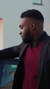 Kırmızı kazaklı kendine güvenen bir adam gösterişli bir arabaya yaslanıyor. Başarıyı somutlaştıran modern bir binanın önünde hareket ediyor.