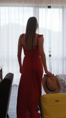 Canlı kırmızı bir elbise giymiş şık bir kadın zarif bir şekilde bavulunun yanında duruyor ve heyecan verici bir yolculuğa hazırlanıyor.