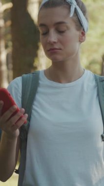 Şık bir sırt çantasıyla genç bir kadın telefonunu dikkatle inceliyor, dingin bir ormanda yemyeşil ağaçlarla çevrili.