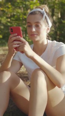 Bir kadın dışarıda oturuyor, akıllı telefonunu kullanıyor ve teknoloji ile doğa arasındaki bağlantıyı vurguluyor.