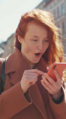 Neşeli kızıl saçlı bir kadın akıllı telefonuyla meşgul oluyor ve mesajlara bakarken neşe saçıyor. Gülüşü sahneyi aydınlatıyor, mutluluğu yansıtıyor.