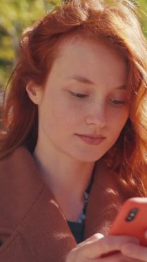 Çarpıcı kızıl saçlı bir kadın dikkatle akıllı telefonuna bakıyor, ekranında görünen her ne ise içine çekiyor.