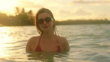 Bikinili bir kadın okyanusa dalmadan önce sığ sularda durup sessizliğin tadını çıkarırken huzurlu bir günbatımı sahnesi gözler önüne seriliyor.