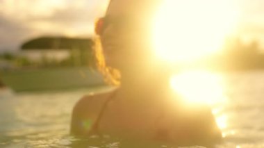 Güneş batarken, bikinili bir kadın su kenarında sakin bir anın tadını çıkarıyor. Sakin okyanusta neşeyle yüzüyor, yazları güzelliği kucaklıyor.