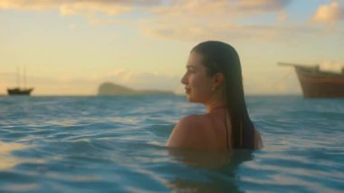 Bir kadın dalgalı okyanusta zarif bir şekilde yüzer, arka planda bir tekne ile. Günbatımı sıcak bir parıltı yayar, özgürlüğü ve doğayı yakalar.