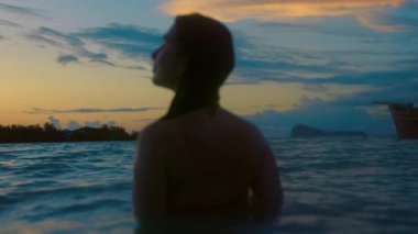 Huzurlu bir günbatımında okyanusta duran bikinili bir kadın görülüyor. Gün batımının ılık renkleri suları yansıtır, sükunet yaratır.