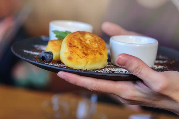 Panquecas de queijo cottage com mirtilo e hortelã para café da manhã ou almoço. Engarrafamento e creme azedo. As mãos da menina segurando o prato acabado . — Fotografia de Stock