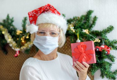 Gülümseyen orta yaşlı, tıbbi maskeli ve Noel Baba şapkalı bir kadın. Noel için uzaktan tebrik ederim. Salgın.