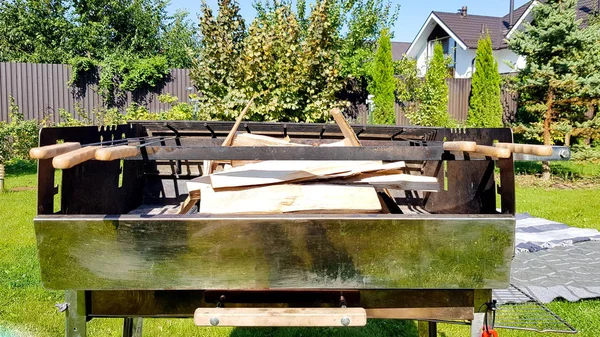 Firewood em um grande churrasco estacionário no quintal da casa, área de churrasco. Preparação para cozinhar carne e legumes na grelha. Piquenique de verão com a família em um gramado verde — Fotografia de Stock