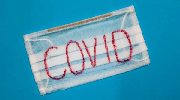 Typical white 3-ply surgical mask with rubber earhooks to cover mouth and nose with English block letters Covid on a blue background. Dangerous virus and quarantine concept