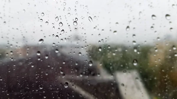 Wet window with drops on the background of the autumn city in cloudy weather. View from the window in the rain. Water drop on glass window during rain with blurred background of city scene.