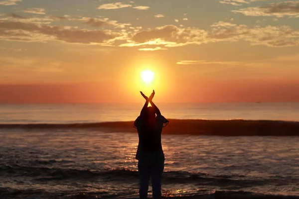 Genç bir kadın silueti ellerini gökyüzüne doğru kaldırıyor ve sabah güneş doğarken deniz kıyısında duruyor. Güneşin ilk parlak ışığını selamlıyor ve teşekkür ediyor.