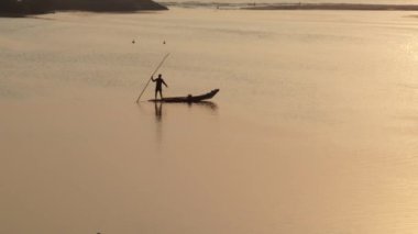 Bir balıkçı silueti gün doğumunda bir teknede oturuyor ve balık yakalamak için ağı nehir suyuna bırakıyor.