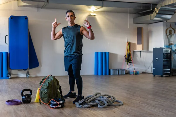 Motivated boot camp instructor stands with gym equipment in gym hall. Dumbbells, rope, sandbag on wooden floor. Instructor showing muscles. Portrait for gym concept.