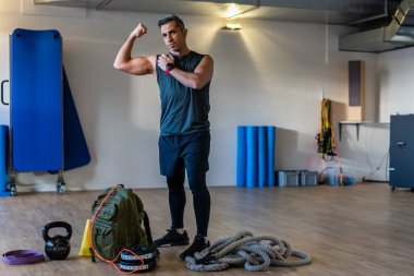 Jimnastik hocası kaslarını gösteriyor. Acemi birliği eğitmeni spor salonundaki spor aletleriyle ayakta duruyor. Dambıllar, ip, tahta zeminde kum torbası. Beden eğitimi konsepti portresi.