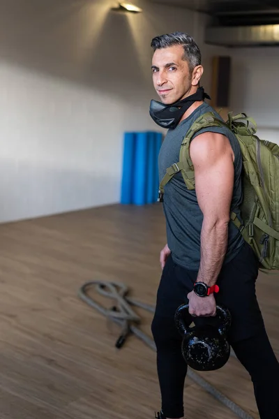 Motivated boot camp instructor stands with gym equipment in gym hall. Carry training mask, dumbbells and sandbag, rope on wooden floor. Instructor smiling to camera. Portrait for gym concept.