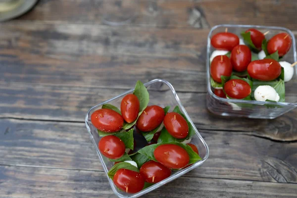 Closeup of red cherry tomatoes with fresh basil and mozzarella cheese. Appetizers sprinkled with salt and pepper, served on small square glass plates on wooden table. High angle view.