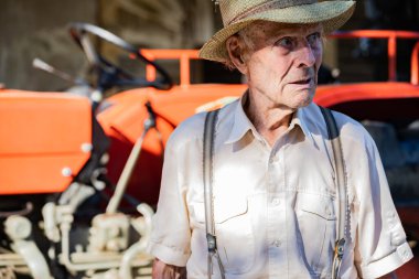 Hasır şapkalı çok yaşlı bir çiftçinin portresi kırmızı bir traktörün önünde hayatı açıklıyor. Gerçek retro, yakın plan..
