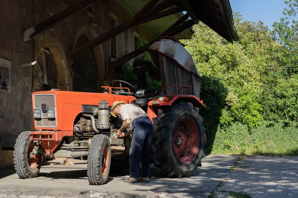 Old farmer working on red tractor under roof of farmer house in sunshine. Vintage, retro, generation, side view.