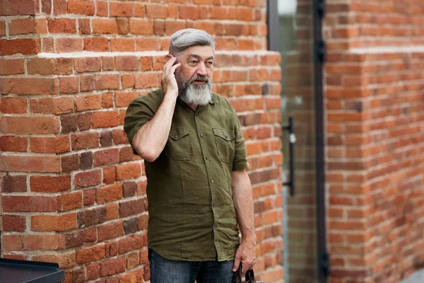 Gray-haired adult businessman hipster in casual clothes standing in the alley of the business center and talking on the phone