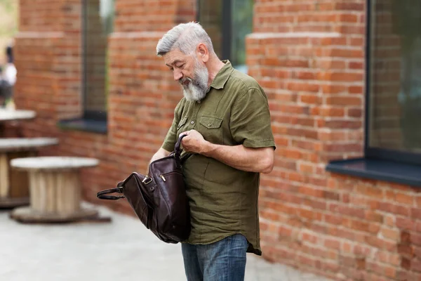 Gray adult businessman hipster in casual clothes standing in an alley downtown and trying to find the documents and the contract in a leather portfolio