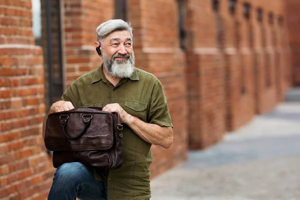 Gray adult businessman hipster in casual clothes standing in an alley downtown and with a smile that looks in a leather bag