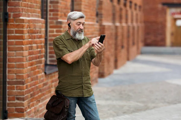 Gray-haired businessman hipster in casual clothes standing in the alley of the business center and dials a message on your mobile phone