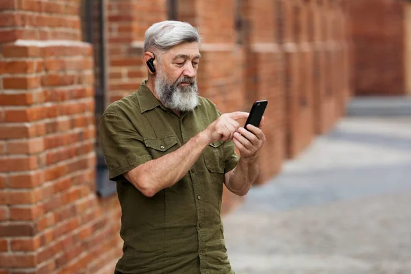 Gray-haired businessman hipster in casual clothes standing in the alley of the business center and dials a message on your mobile phone